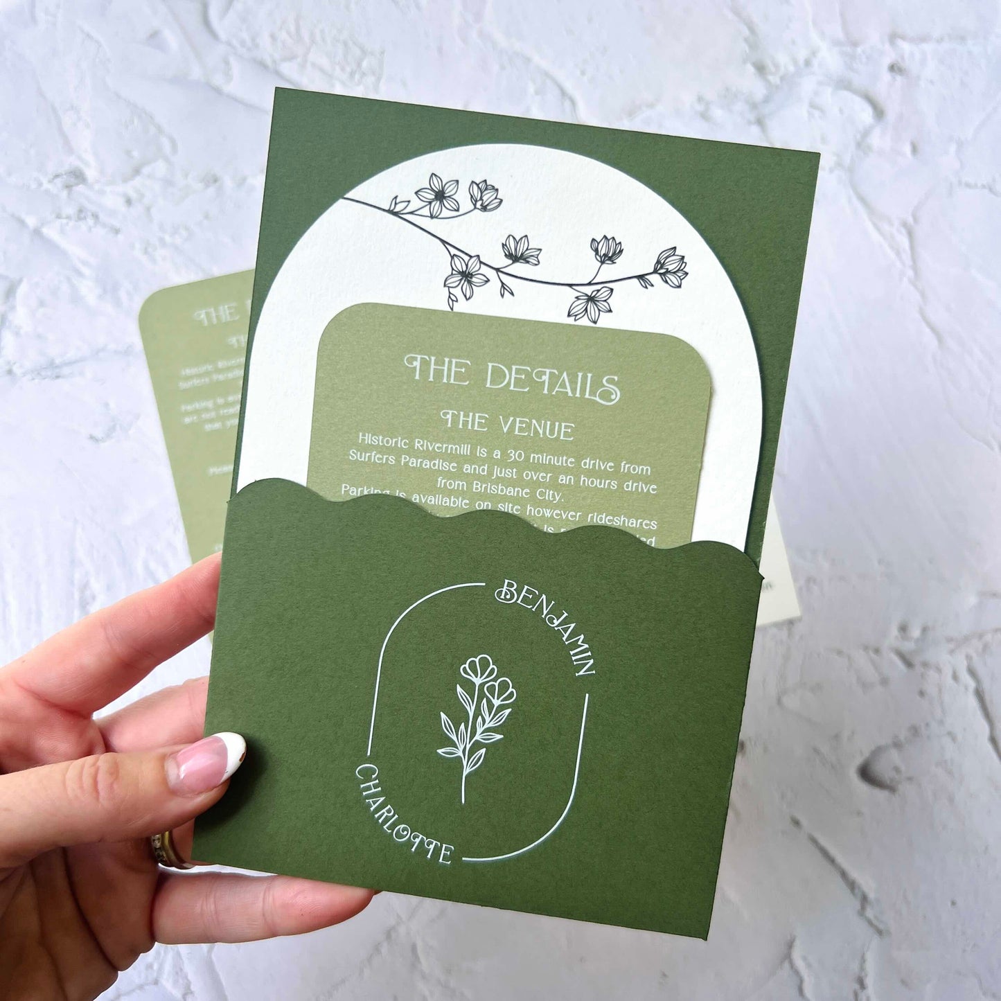 Green invitation card with floral design held by a hand on a light gray background