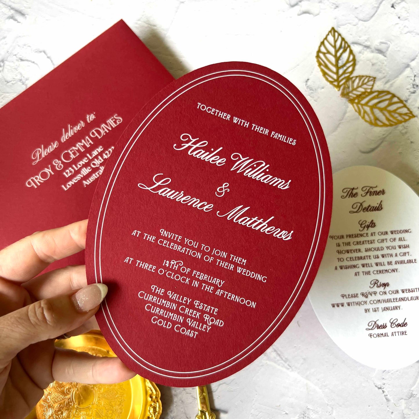 Red wedding invitation card held by a hand with gold leaves in the background
