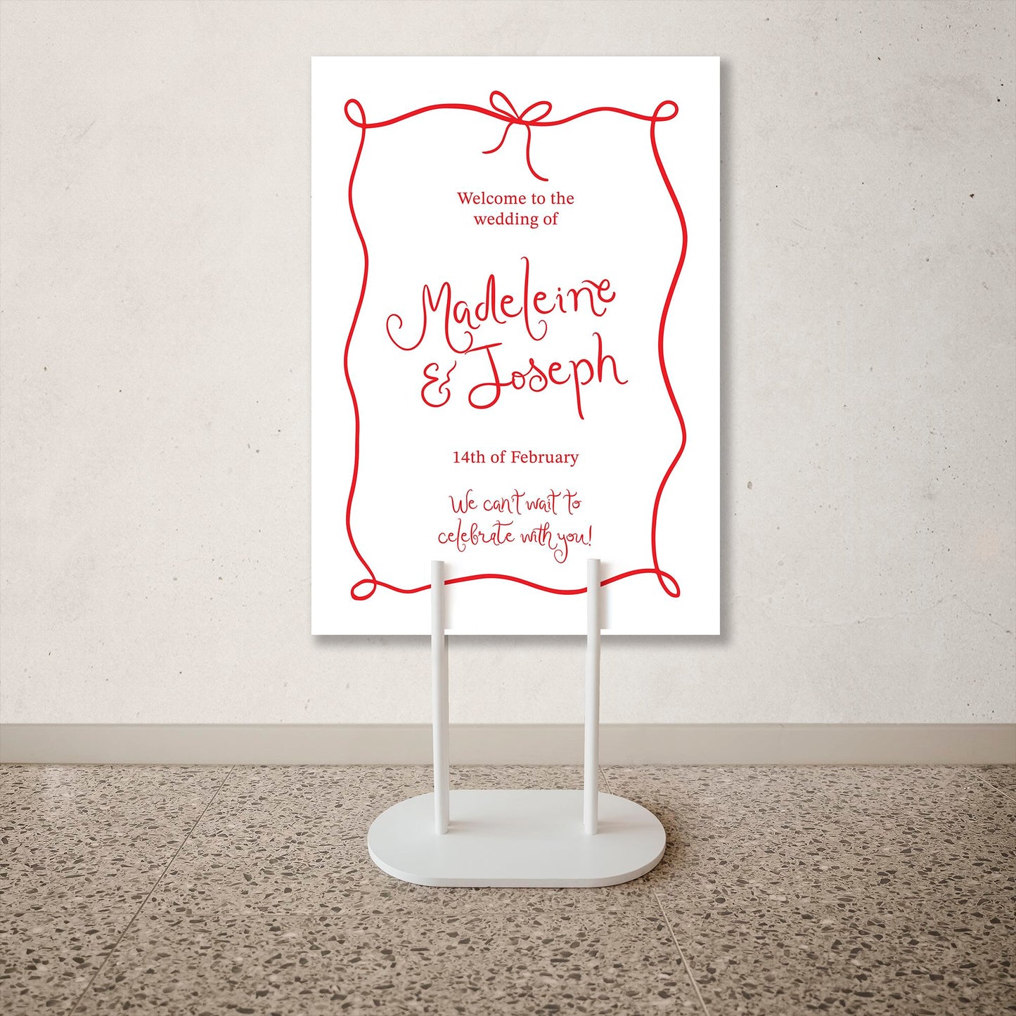 Wedding welcome sign with red text on a white background, displayed on a stand.
