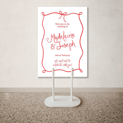 Wedding welcome sign with red text on a white background, displayed on a stand.