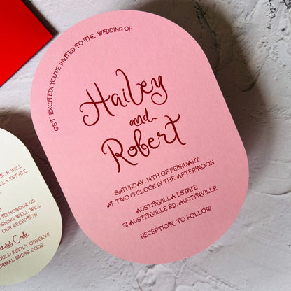 Pink oval wedding invitation card with red text on a textured surface