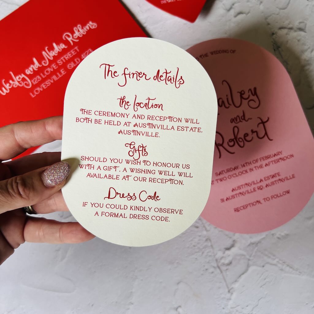 Oval wedding invitation card with red text held by a hand on a light background