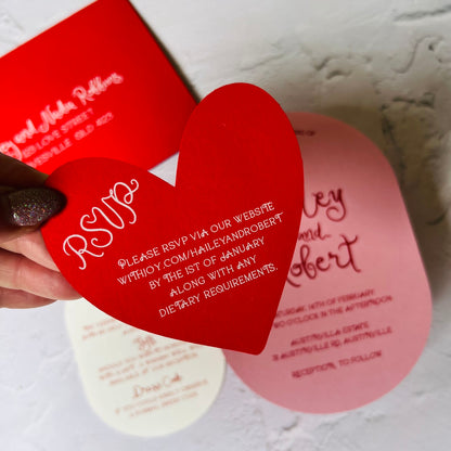 Red heart-shaped RSVP card held over a wedding invitation with text.