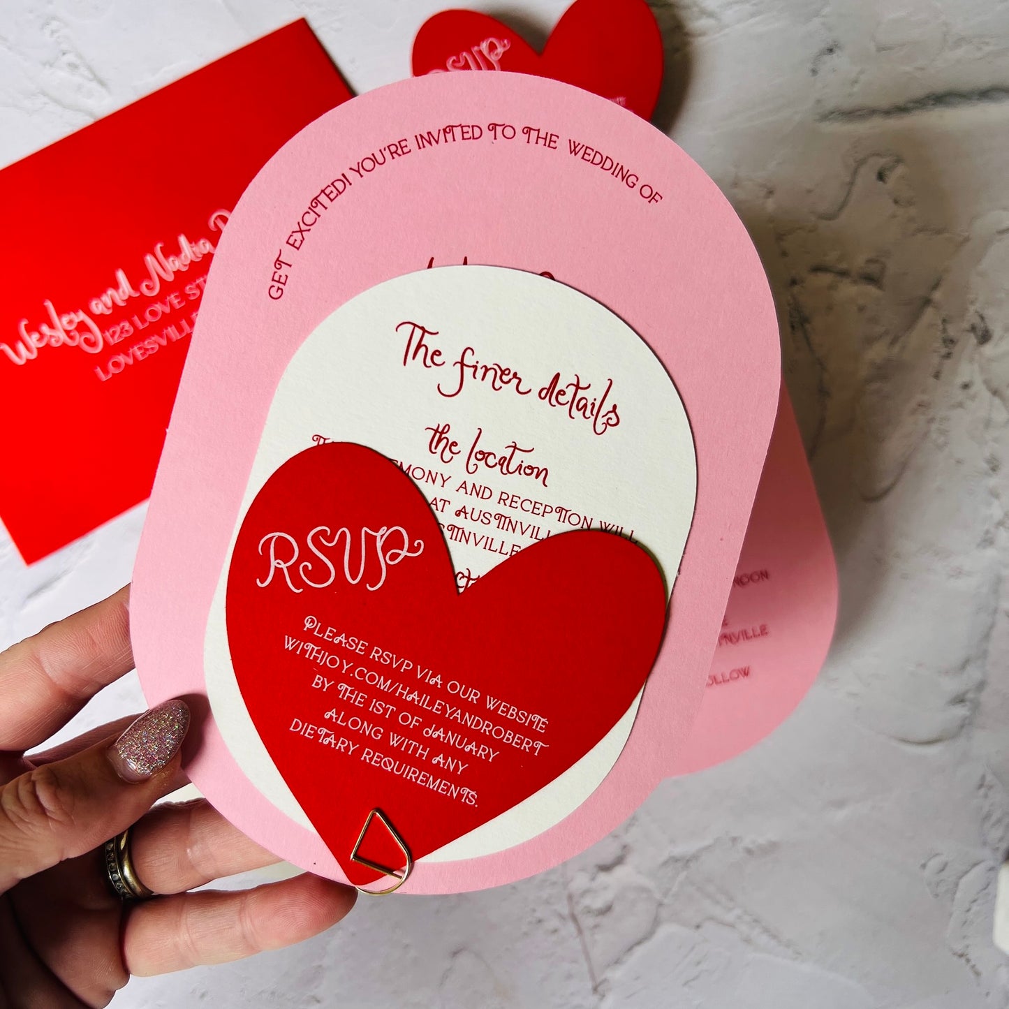 Heart-shaped and oval wedding invitation with red and pink colors, held by a hand.