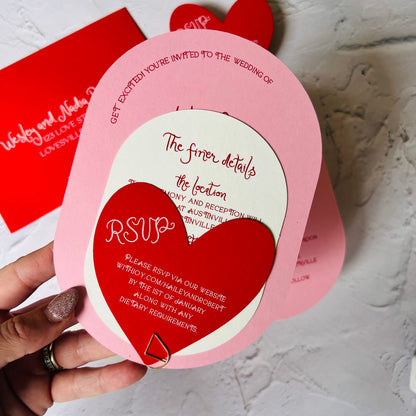 Heart-shaped and oval wedding invitation with red and pink colors, held by a hand.