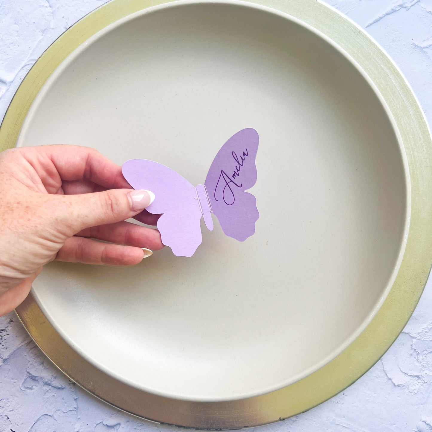 Hand holding a wedding purple butterfly-shaped card with 'Anelia' on it, over a beige plate.
