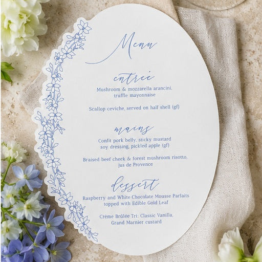 Oval menu card with blue text and decorative border on a beige surface with flowers
