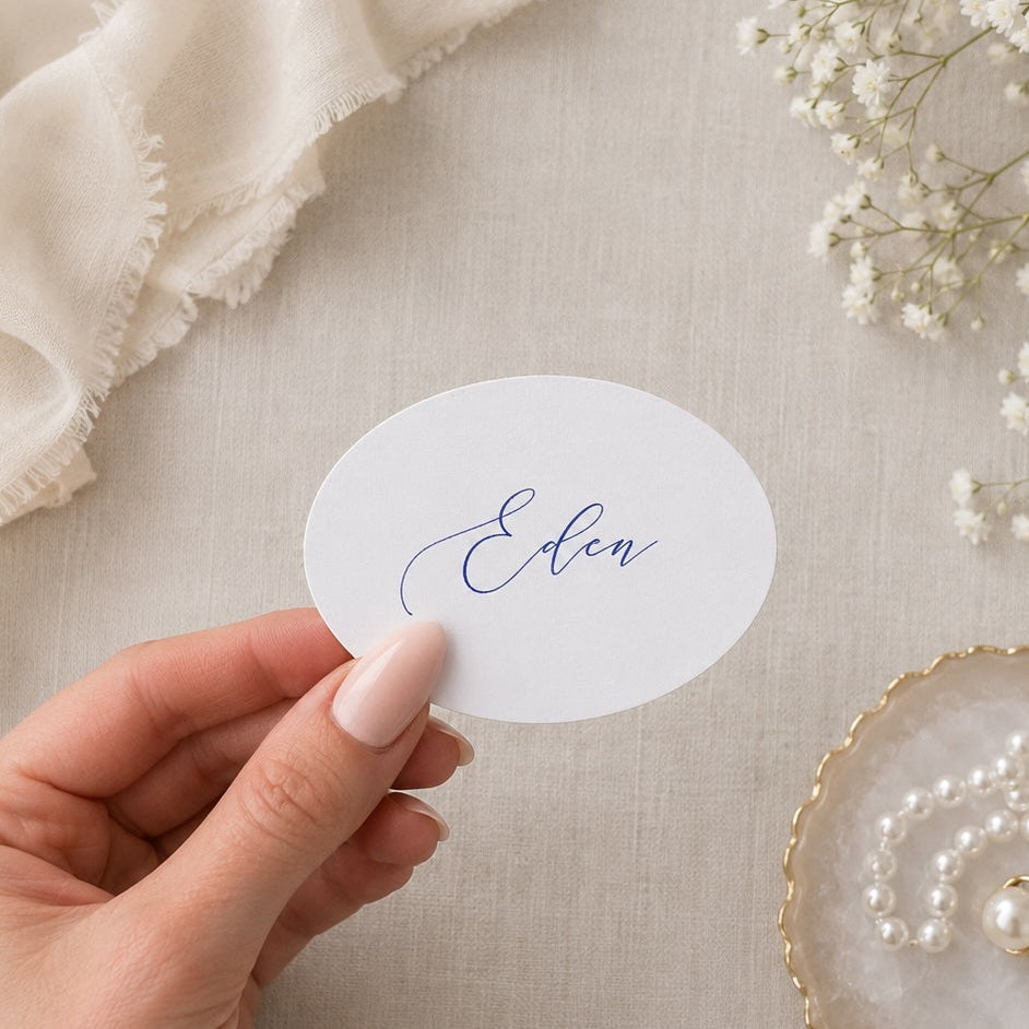 Hand holding a round card with 'Eden' written on it, surrounded by white flowers and jewelry on a light fabric background.