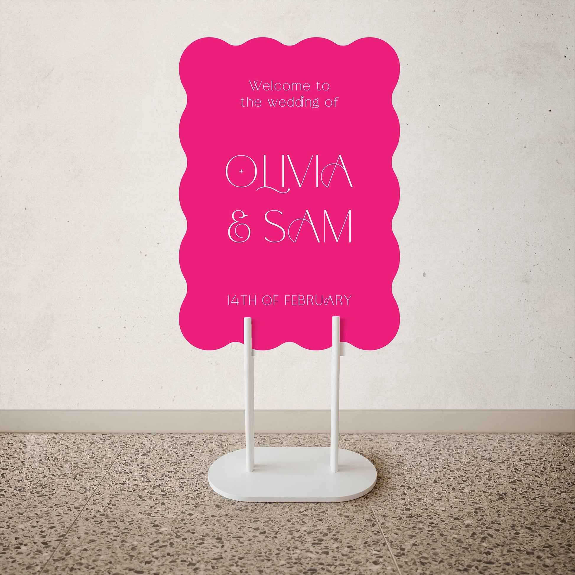 Wave Shape Wedding Welcome Sign Pink wedding sign with white text on a stand against a light gray wall.