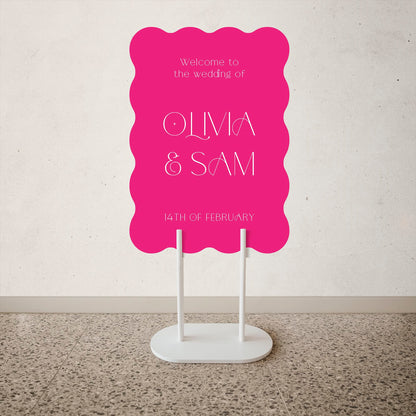 Wave Shape Wedding Welcome Sign Pink wedding sign with white text on a stand against a light gray wall.
