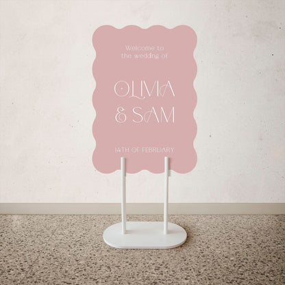 Wave Shape Wedding Welcome Sign  Pink wedding sign with white text on a stand against a light gray wall.