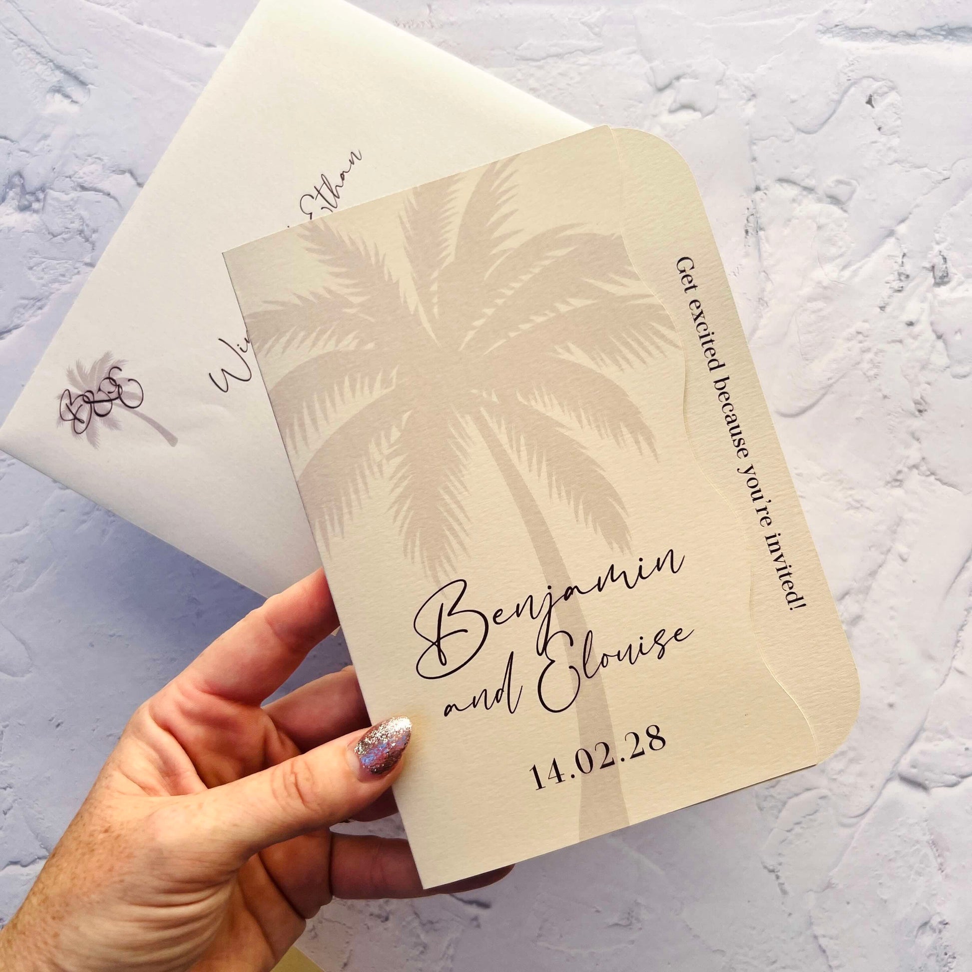 Hand holding a wedding invitation with palm tree design and text on a textured surface