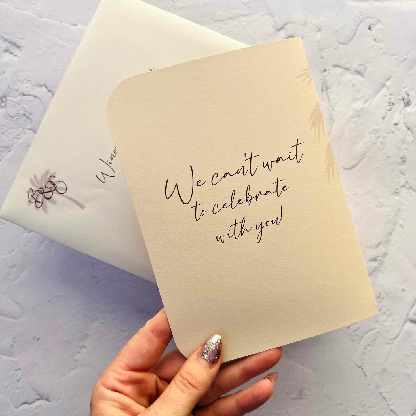 Beige card with 'We can't wait to celebrate with you!' text held by a hand on a light gray background