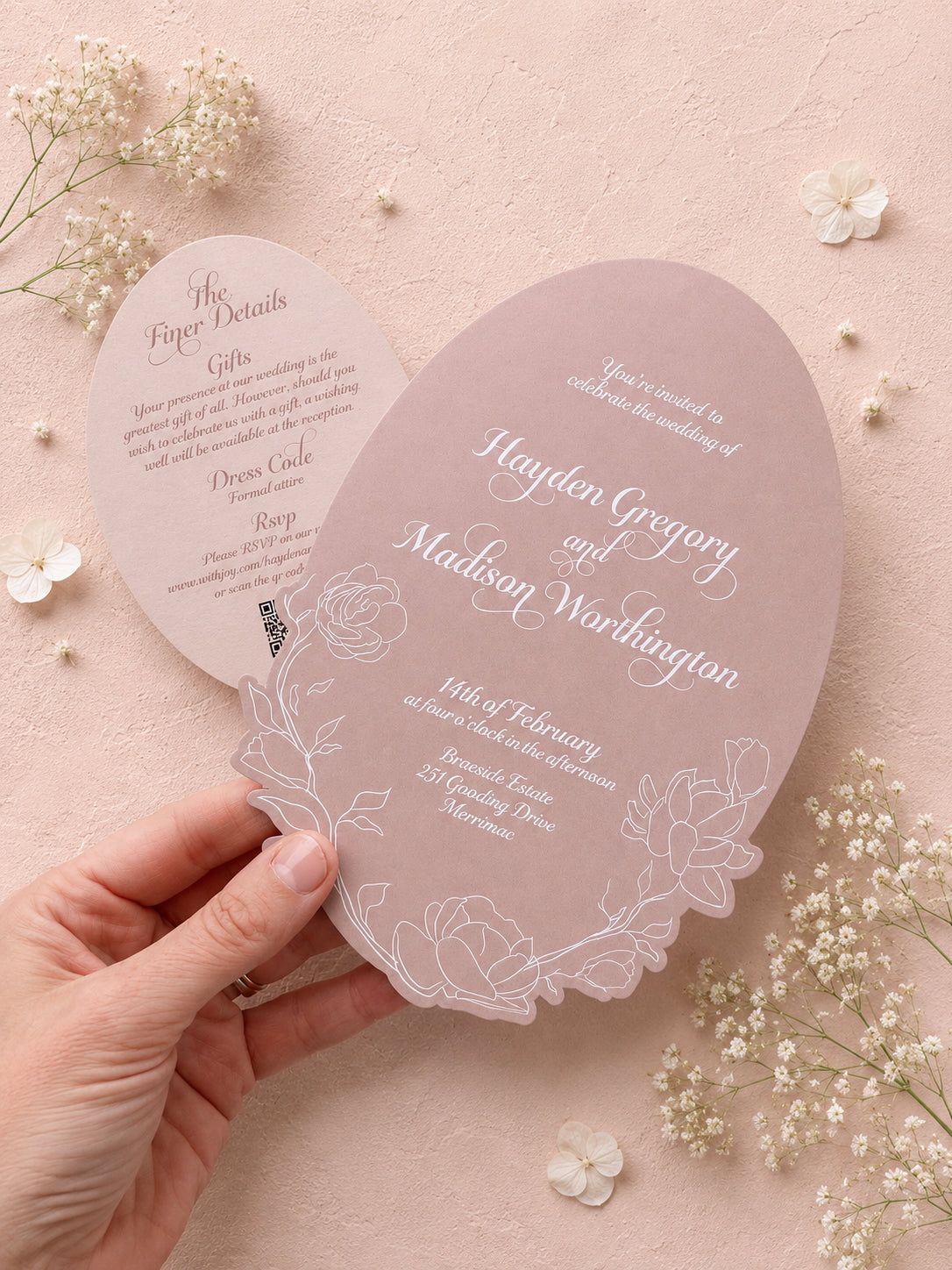 Elegant wedding invitation card held by a hand with floral decorations on a soft pink background